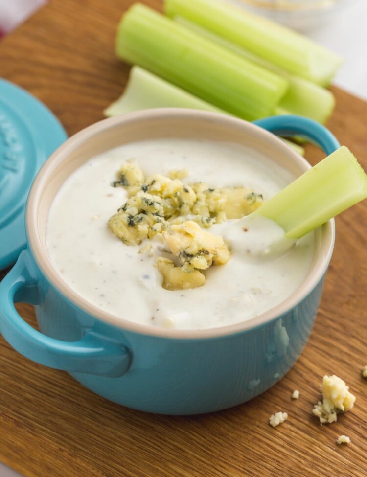 a blue crock filled with blue cheese dressing. a stack of celery sticks is in the background, and one celery stick is in the blue cheese.