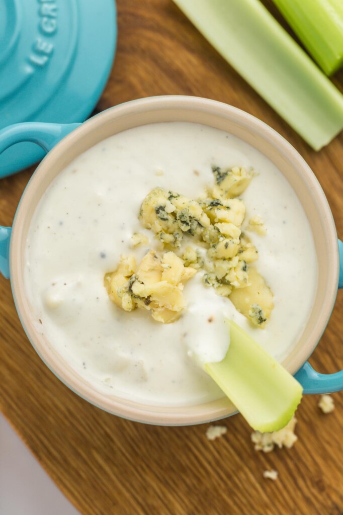 a crock of of blue cheese dressing, viewed from above, with crumbled blue cheese on top and a celery stick stuck in.