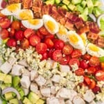 a large white platter of chicken cobb salad made with layers of avocado, chicken, tomato, egg, bacon, and lettuce