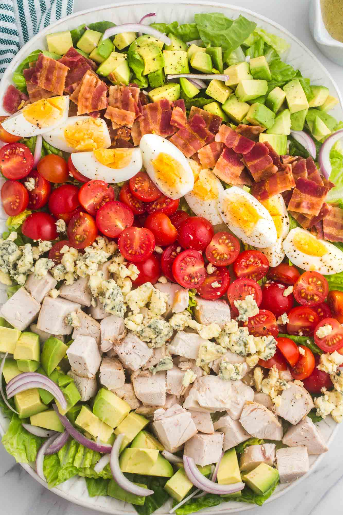 a large white platter of chicken cobb salad made with layers of avocado, chicken, tomato, egg, bacon, and lettuce