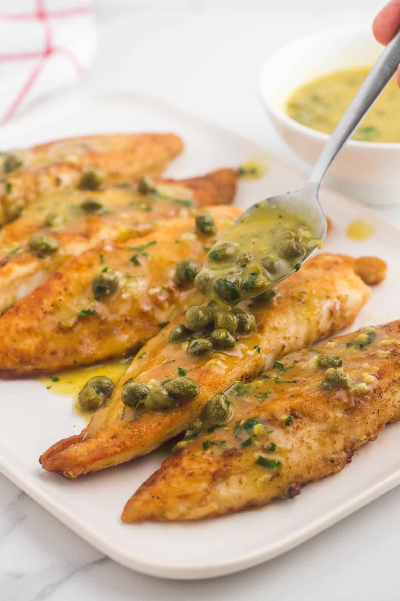 a platter of browned fish fillets being topped with capers piccata sauce