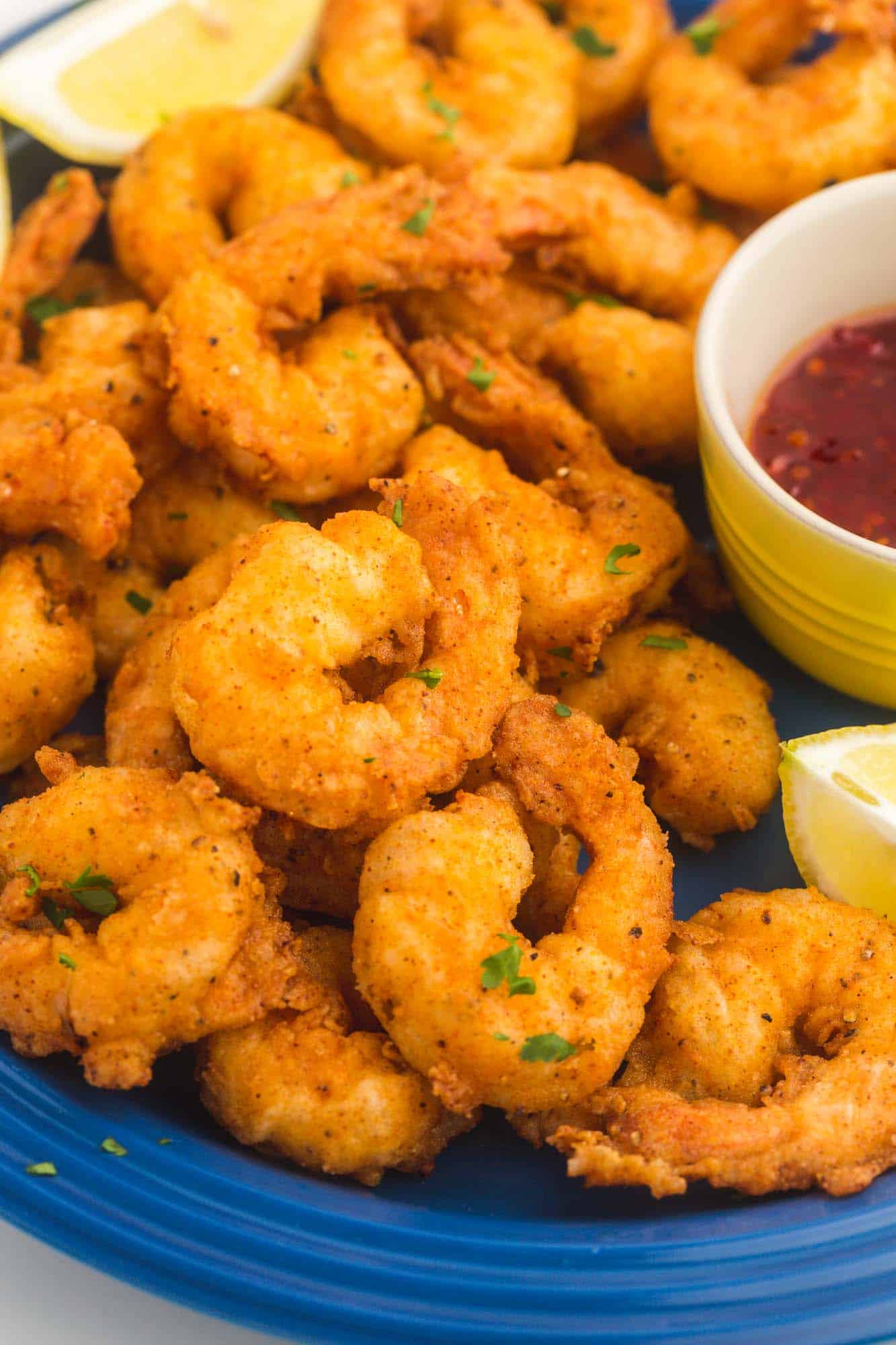 cropped image of a blue plate filled with fried shrimp. Lemon wedges garnish the plate and a cup of dipping sauce is on the side