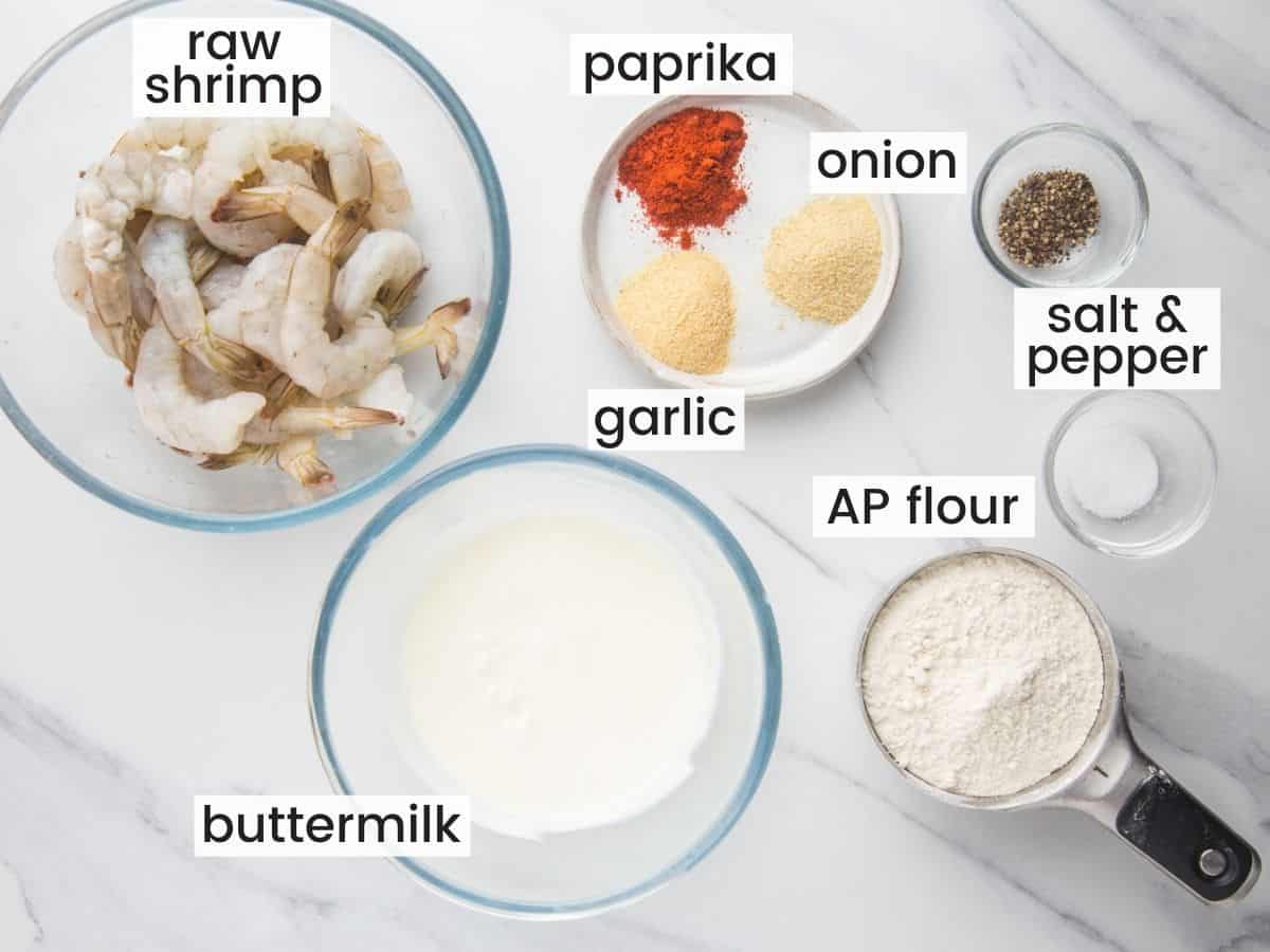 The ingredients needed to make fried shrimp, measured out into separate bowls on a marble countertop, viewed from above