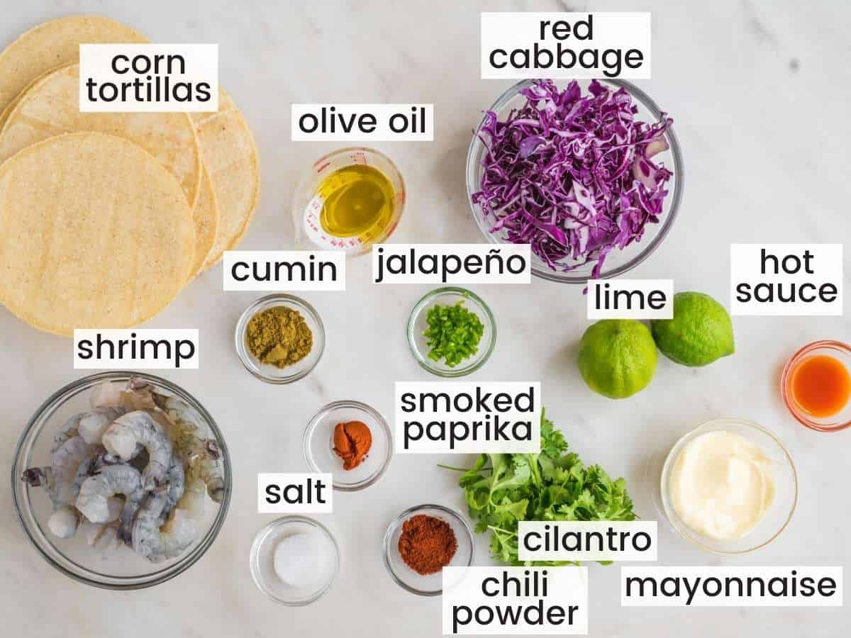 Ingredients needed for making shrimp tacos including shrimp, spices, lime, cilantro, mayo, hot sauce, corn tortillas, cilantro and olive oil.
