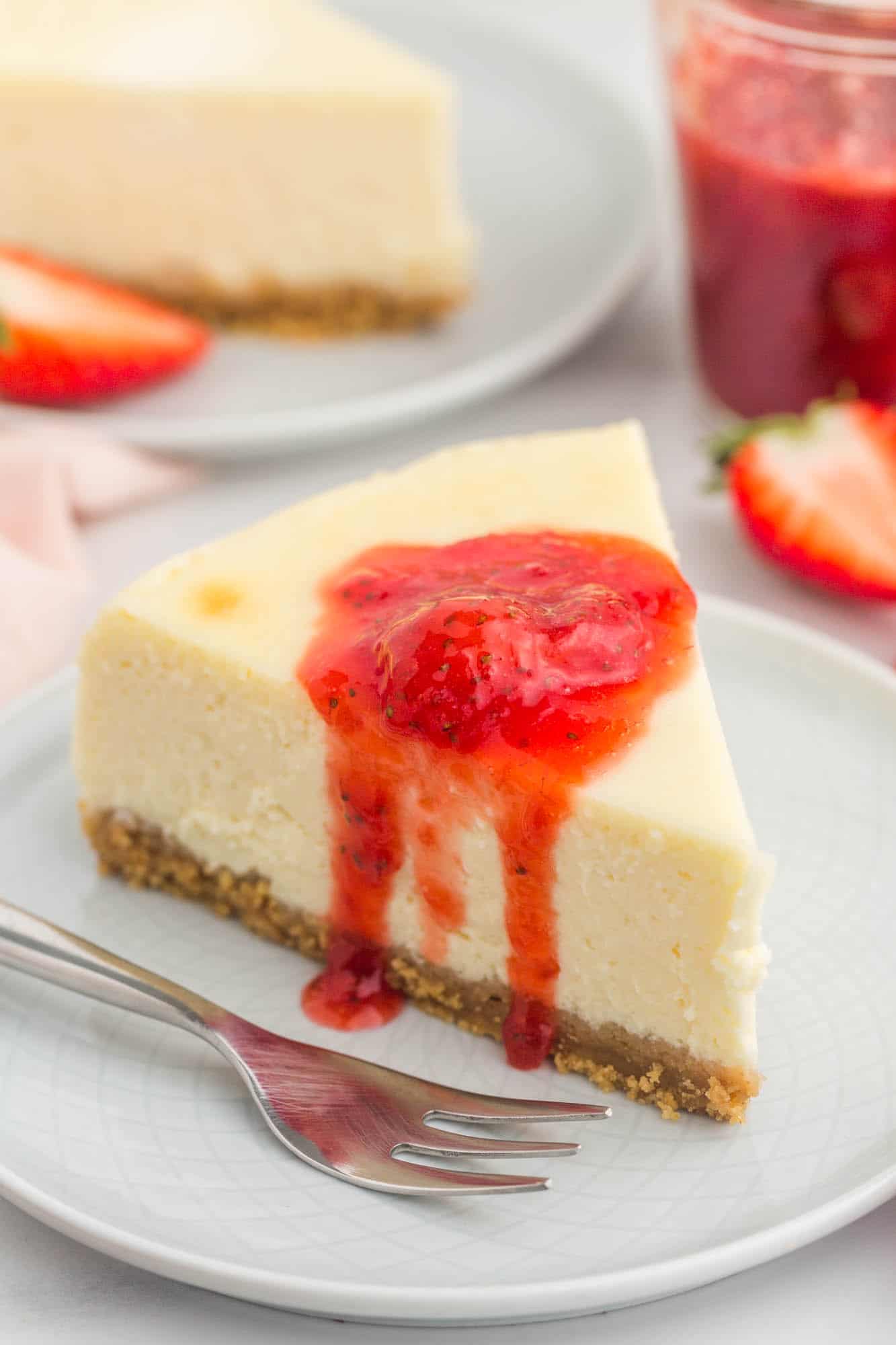 a slice of NY style cheesecake on a small white plate. It's topped with a dollop of strawberry sauce. 