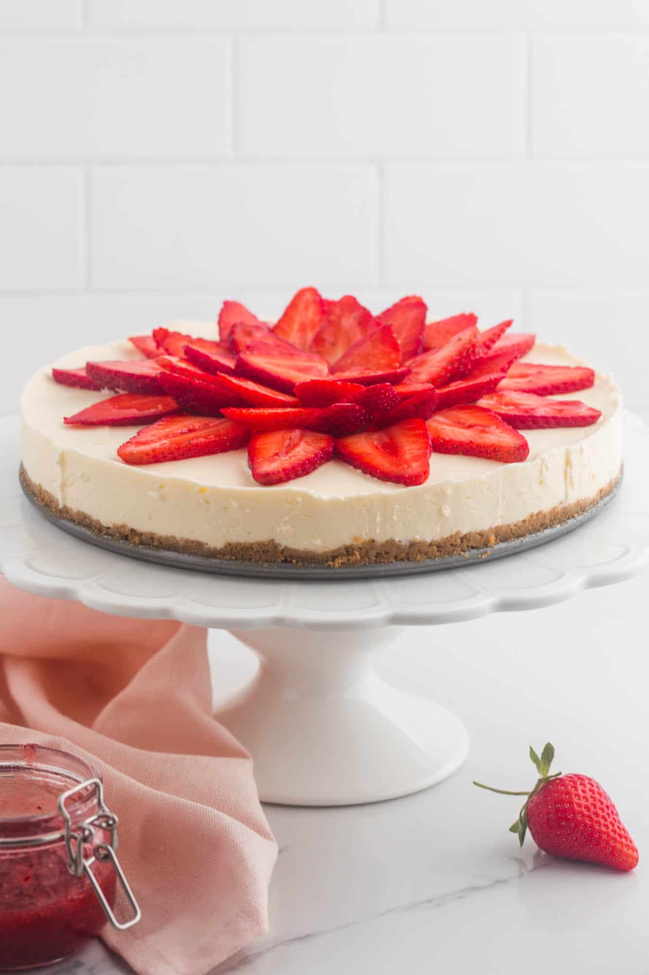 a white cake pedestal holding a no bake cheesecake topped with sliced strawberries that look like a lotus flower