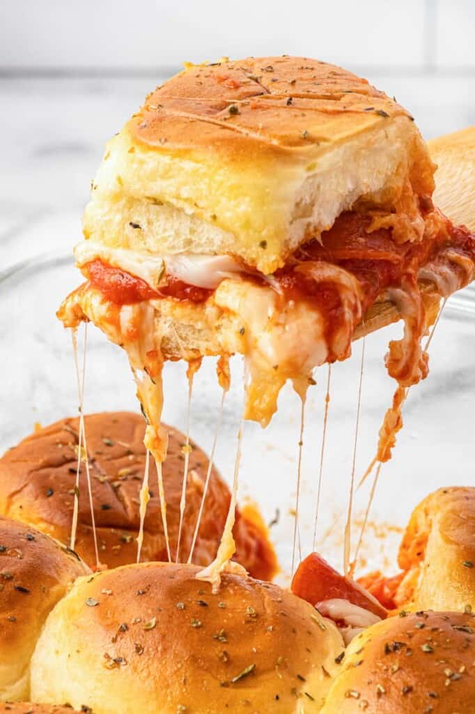 a pizza slider on a Hawaiian roll being lifted out of the pan, mozzarella cheese strings hang down.