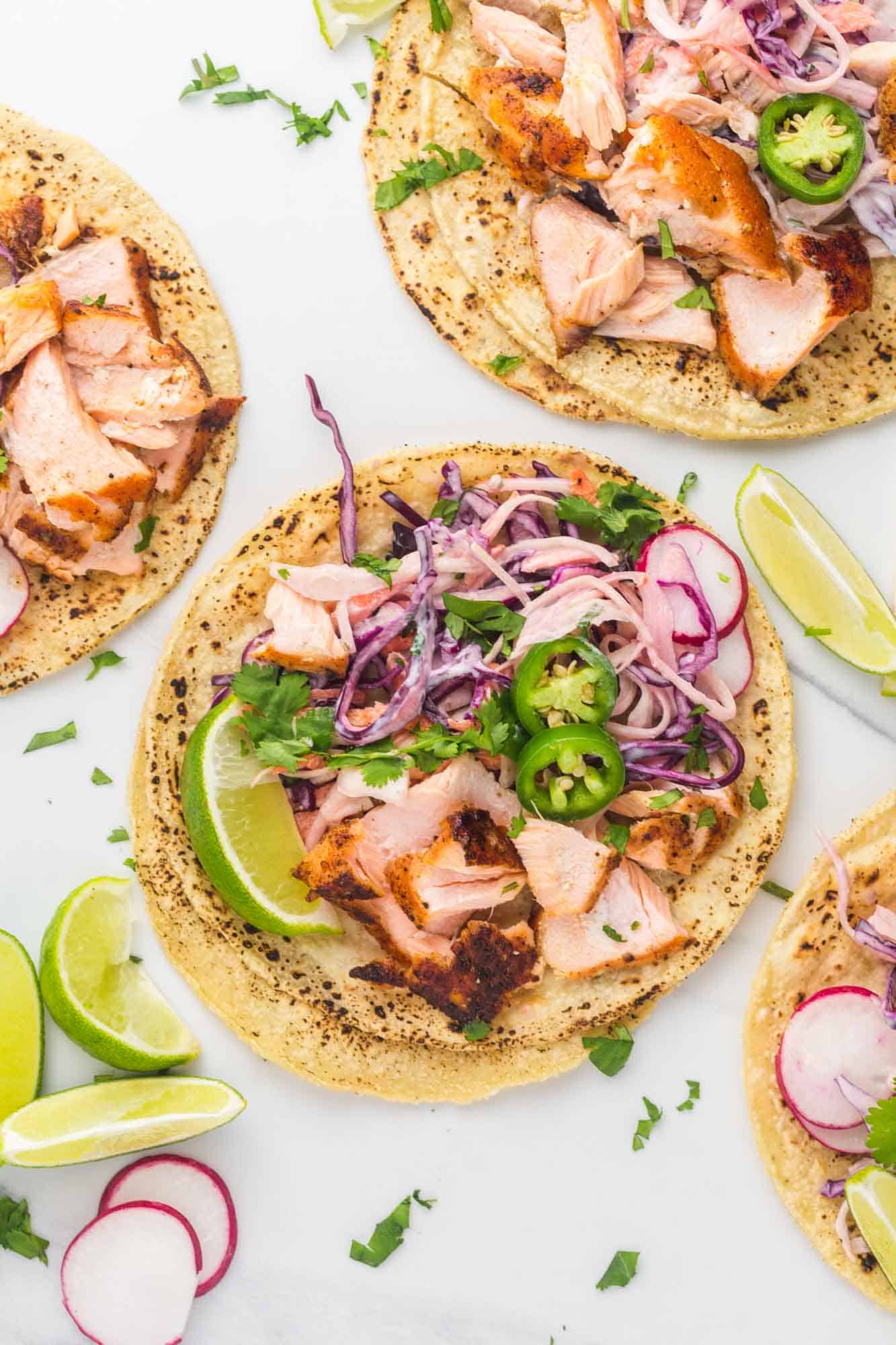 a corn tortilla topped with flaked salmon and fish taco slaw, sliced radishes and jalapenos, and a lime wedge.