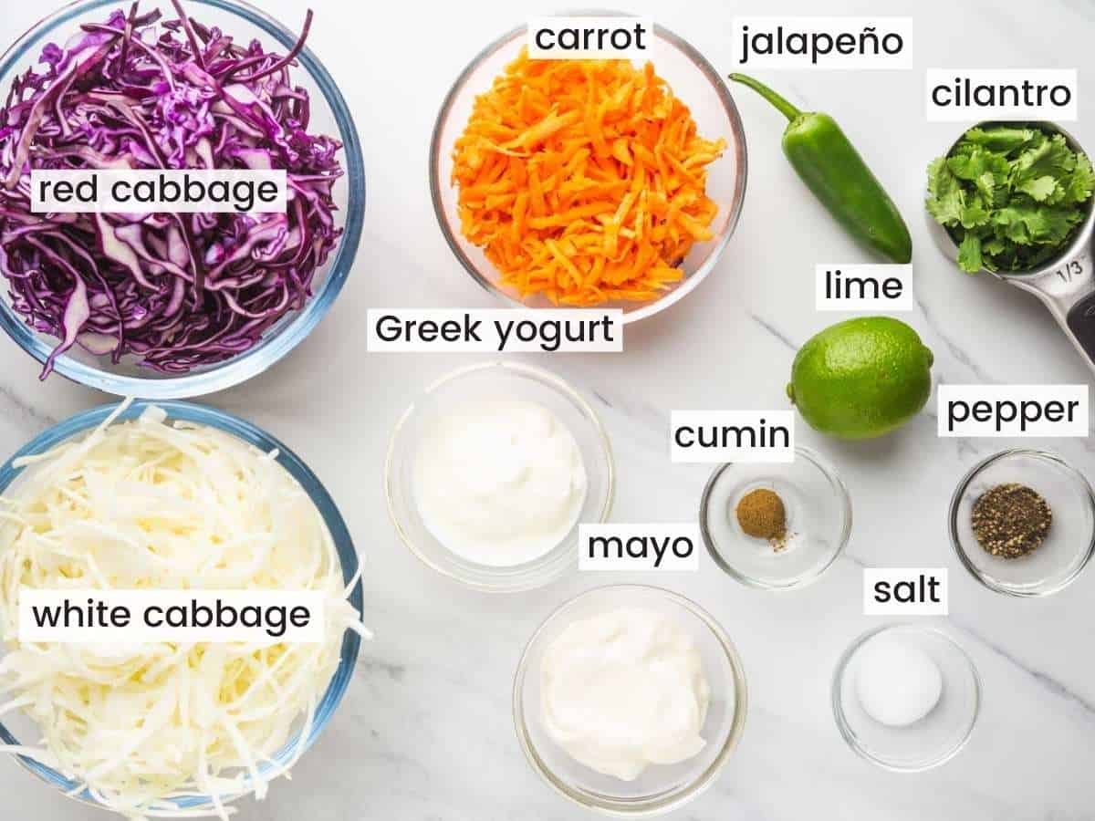 The ingredients for fish taco slaw in separate bowls on a counter. Includes white and red cabbage, carrots, jalapeno, lime, and other ingredients