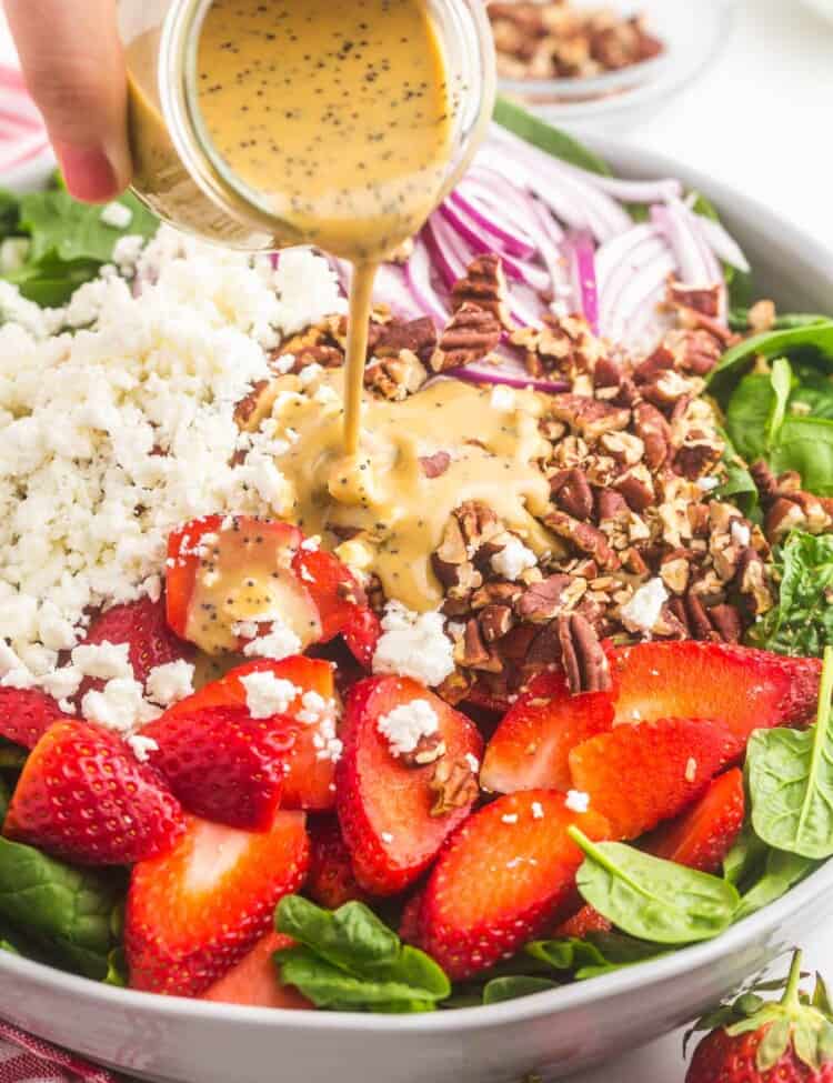 A bowl of spinach salad topped with strawberries, nuts, and cheese. Balsamic dressing is being poured on top