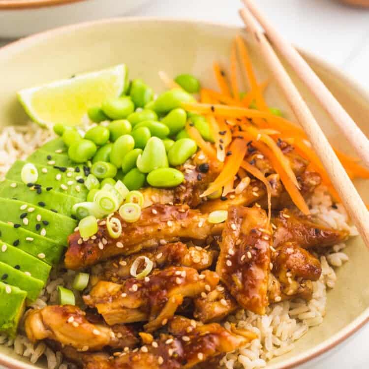 cream colored bowl filled with rice, topped with teriyaki chicken and vegetables. Chopsticks rest on the right side of the bowl, a lime wedges is on the left side