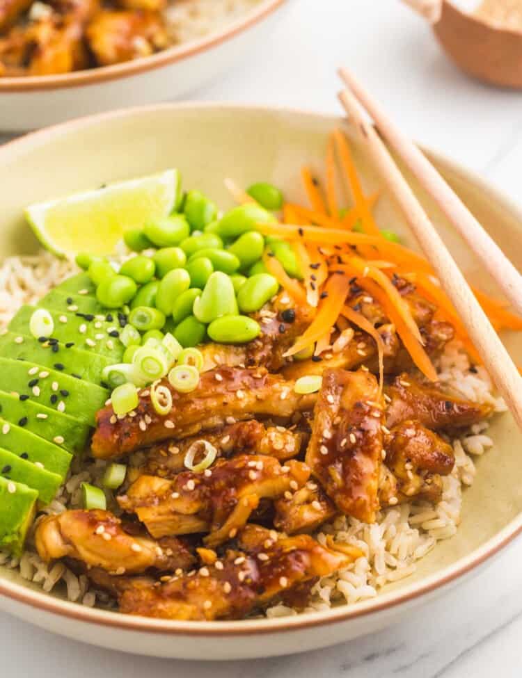 cream colored bowl filled with rice, topped with teriyaki chicken and vegetables. Chopsticks rest on the right side of the bowl, a lime wedges is on the left side