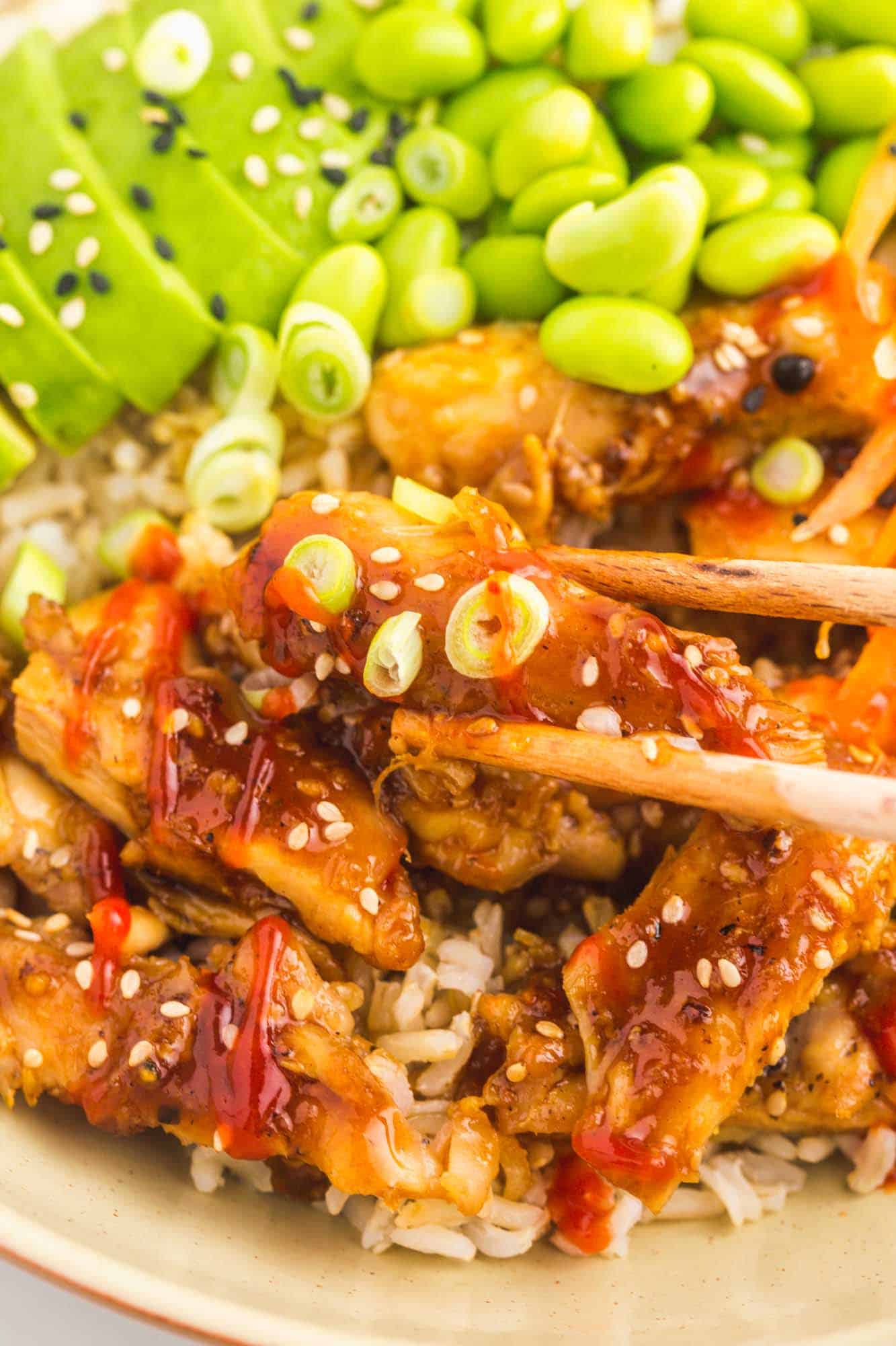 closup view of a bowl of rice, teriyaki chicken, and edamame being eaten with chopsticks.
