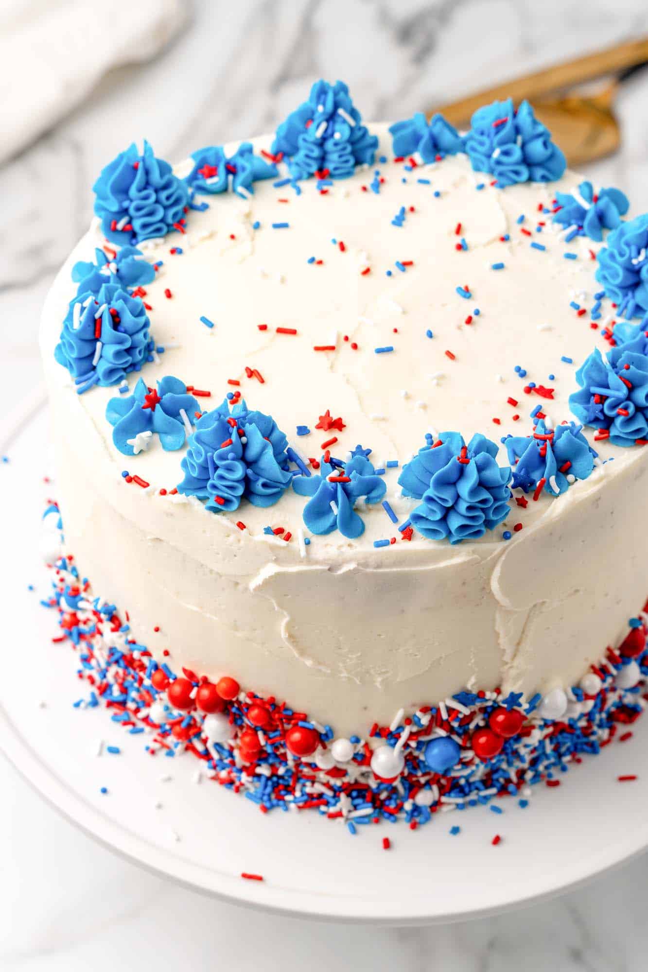 a layer cake with white frosting, a blue top border, and red white ad blue sprinkles on top and piled at the base. 