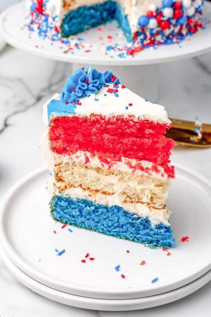 A triangular slice of a red white and blue layer cake with buttercream frosting and colorful sprinkles.