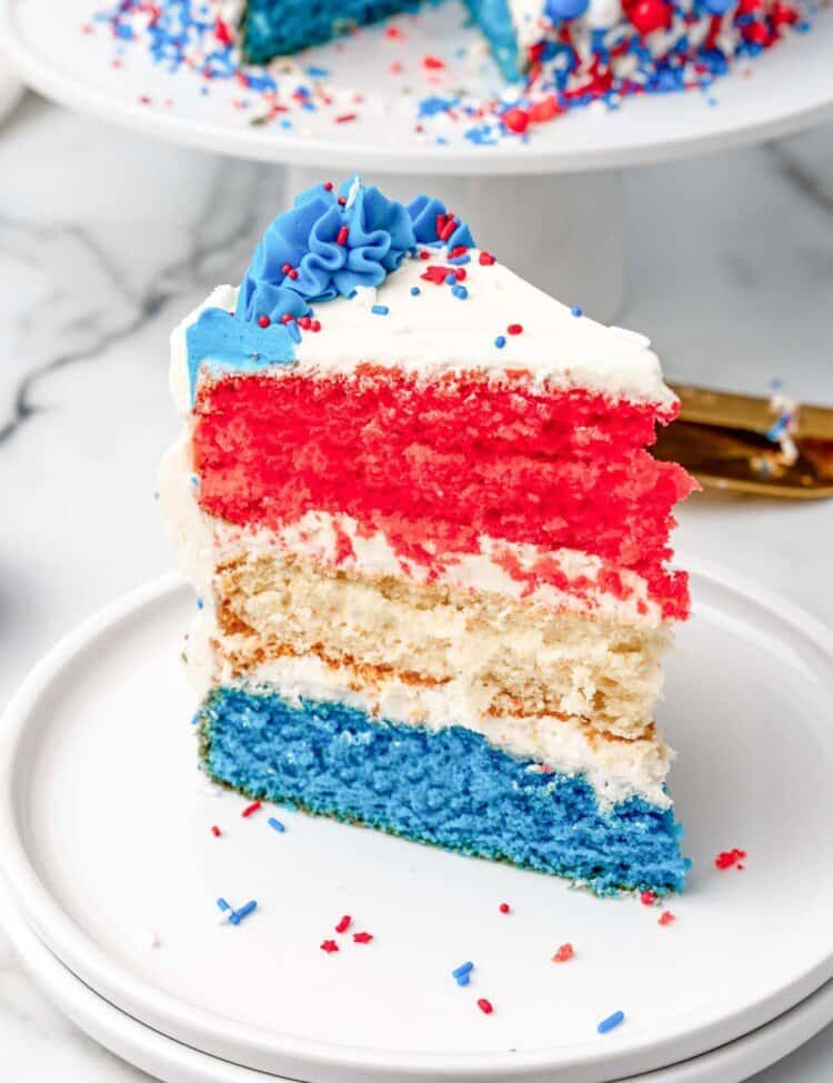 A triangular slice of a red white and blue layer cake with buttercream frosting and colorful sprinkles.