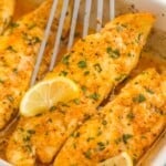 a casserole dish of baked tilapia fillets, garnished with lemon wedges. A fish flipper is lifting up a fillet.