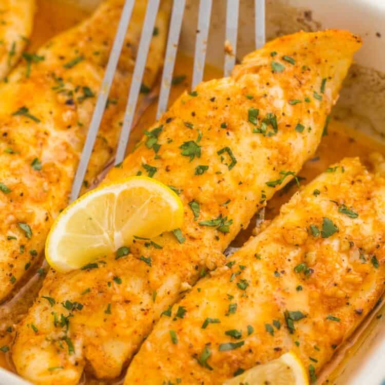a casserole dish of baked tilapia fillets, garnished with lemon wedges. A fish flipper is lifting up a fillet.