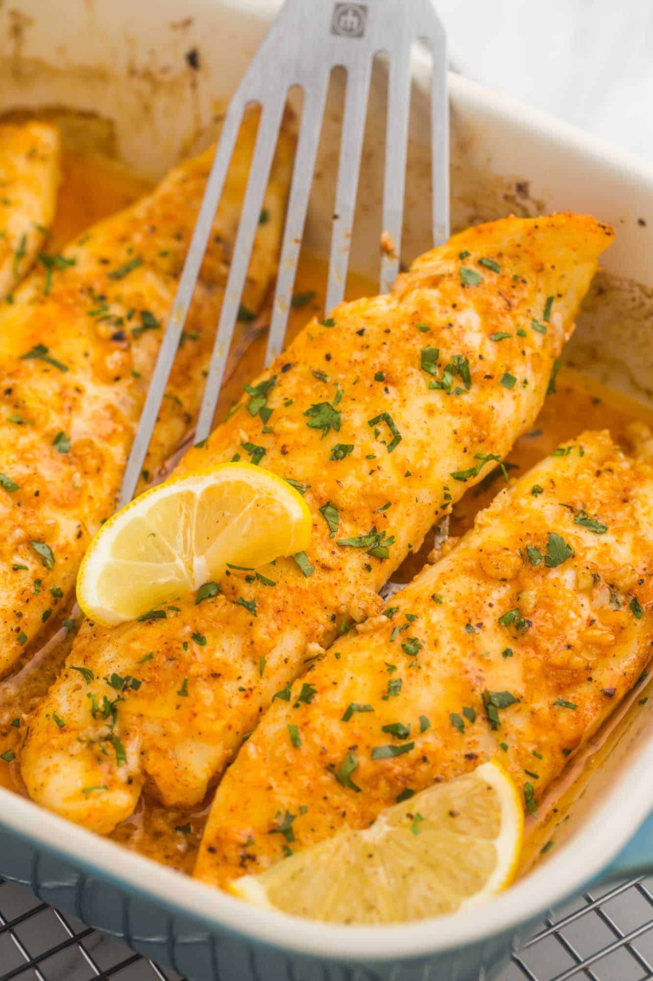 a casserole dish of baked tilapia fillets, garnished with lemon wedges. A fish flipper is lifting up a fillet. 