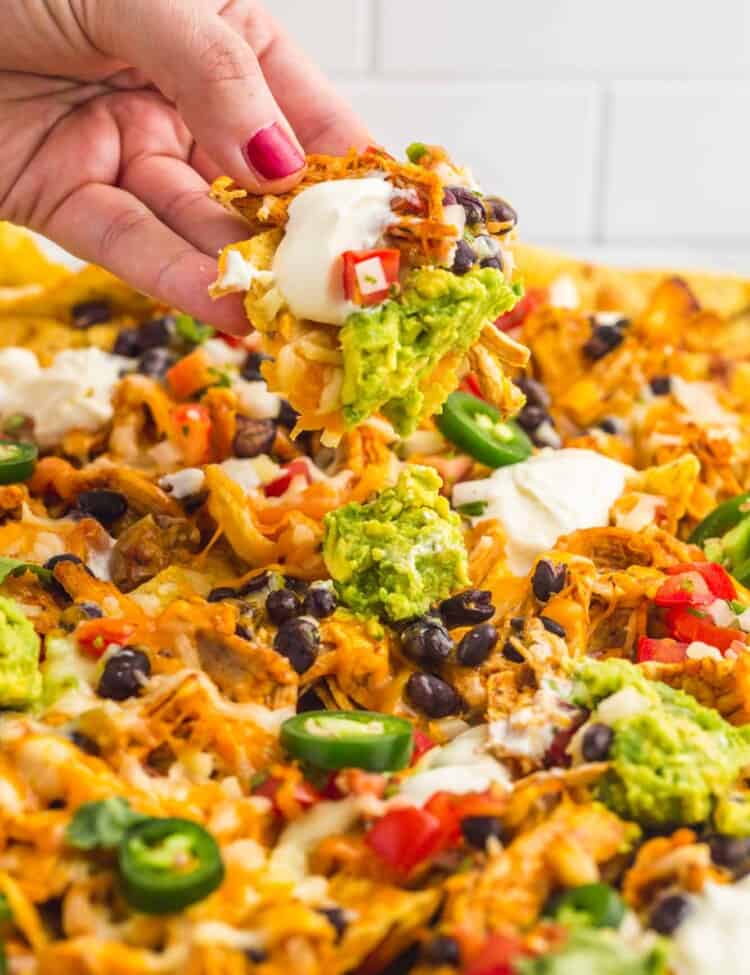 a large tray of nachos topped with chicken, guacamole, sour cream, and jalapenos. A hand is picking up a loaded chip.