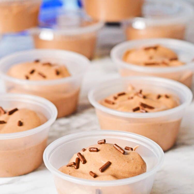 5 open chocolate pudding shots on a counter. In the background is several more shots, stacked up with lids on them.