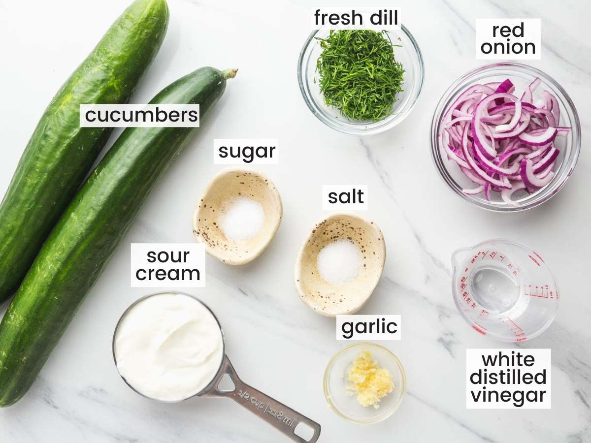 The ingredients for creamy cucumber salad with sour cream, in separate bowls on a marble counter. viewed from above.