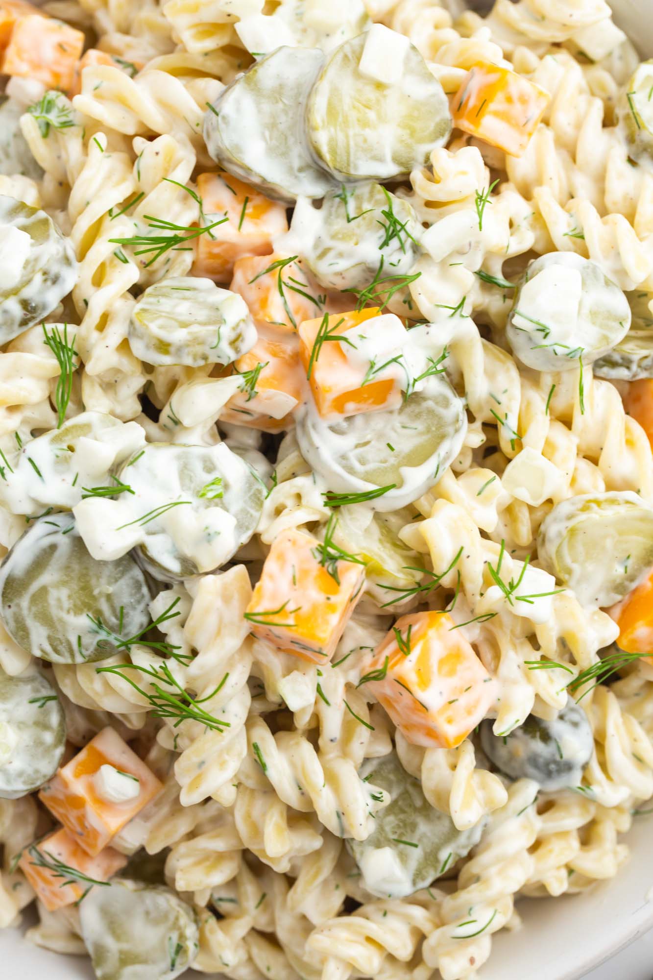 a bowl of dill pickle pasta salad with rotini, cubed cheese, and sliced pickles,The salad is garnished with fresh dill.