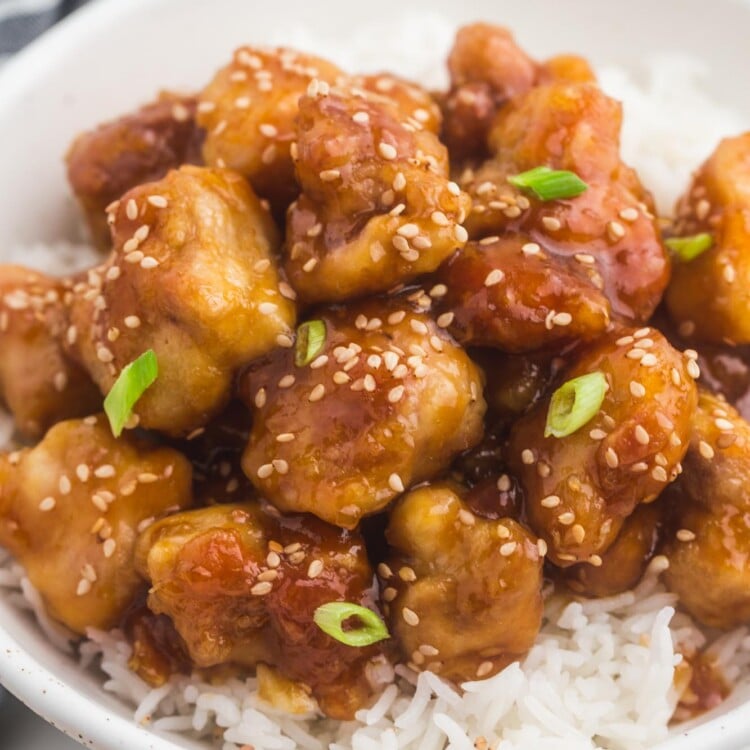 a shallow white bowl filled with jasmine rice and homemade general tso's chicken, garnished with sesame seeds and sliced green onion