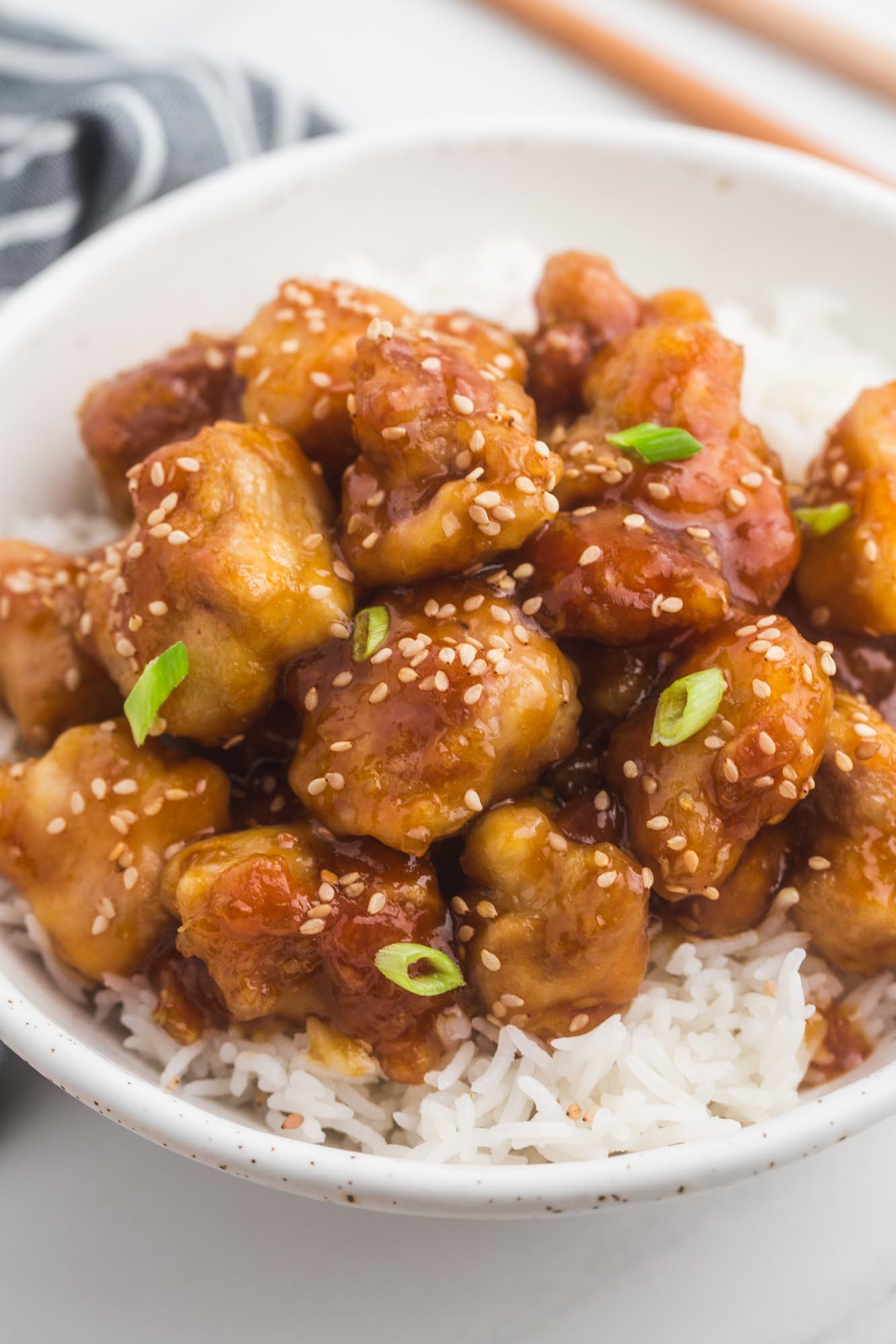 a shallow white bowl filled with jasmine rice and homemade general tso's chicken, garnished with sesame seeds and sliced green onion
