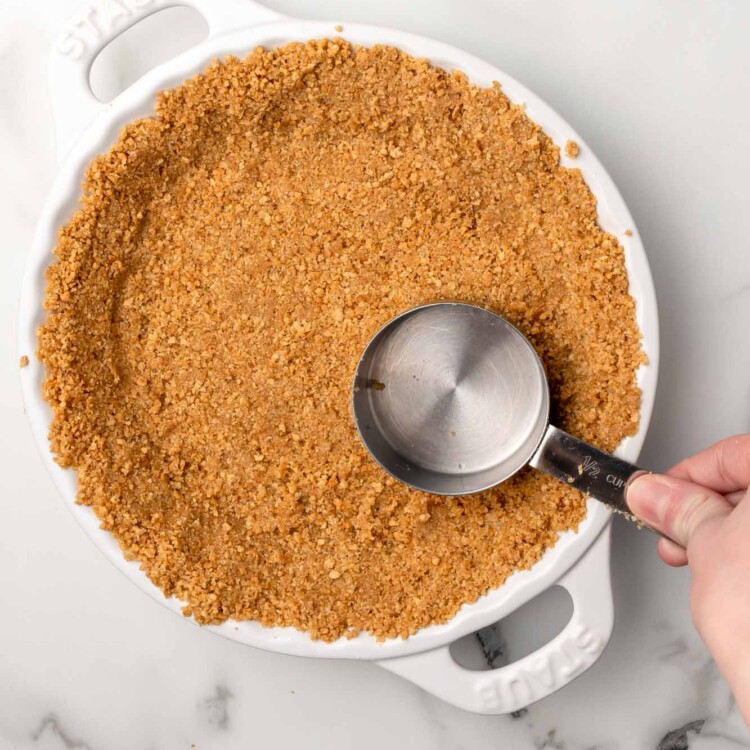 graham cracker crust being pressed into a white ceramic pie plate with a measuring cup