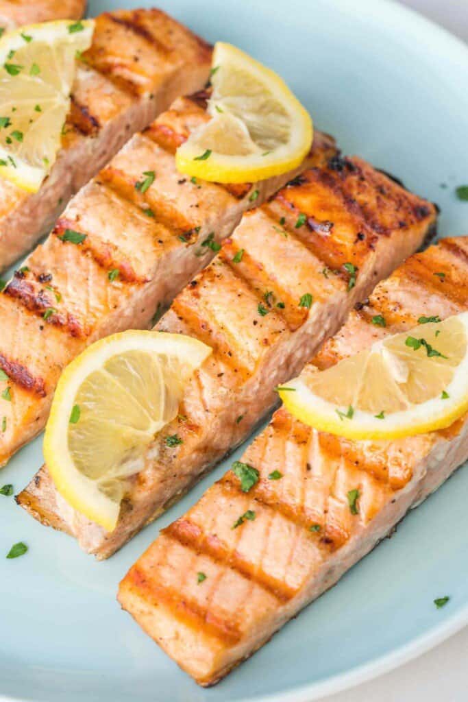 Salmon filets with pronounced grill marks, topped with lemon slices, and resting on a light blue plate.
