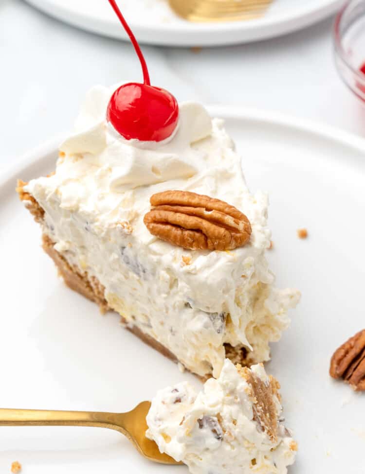 A slice of million dollar pie topped with a maraschino cherry and half a pecan, on a white plate with a bite taken by a gold fork.