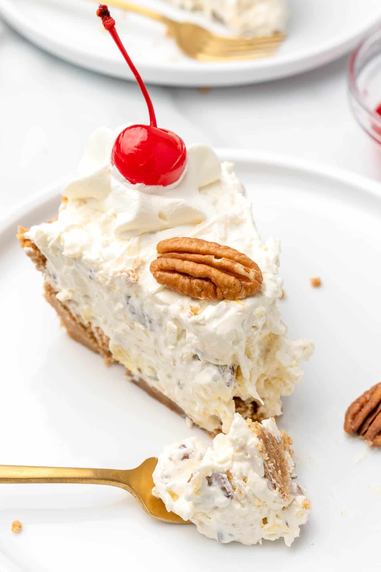 A slice of million dollar pie topped with a maraschino cherry and half a pecan, on a white plate with a bite taken by a gold fork.