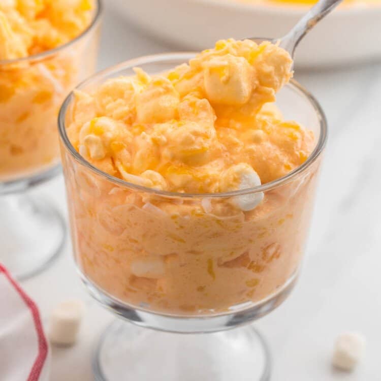 a footed dessert dish filled with a serving of orange fluff jello salad. It's being enjoyed with a spoon.