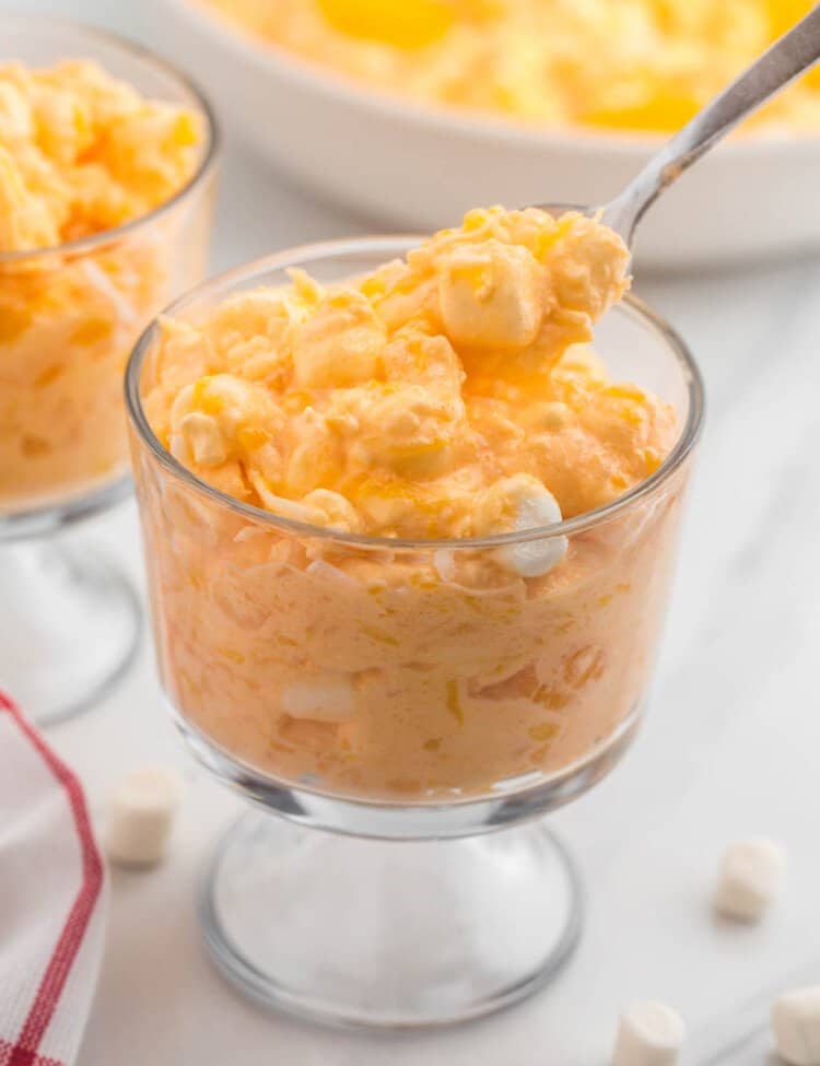 a footed dessert dish filled with a serving of orange fluff jello salad. It's being enjoyed with a spoon.