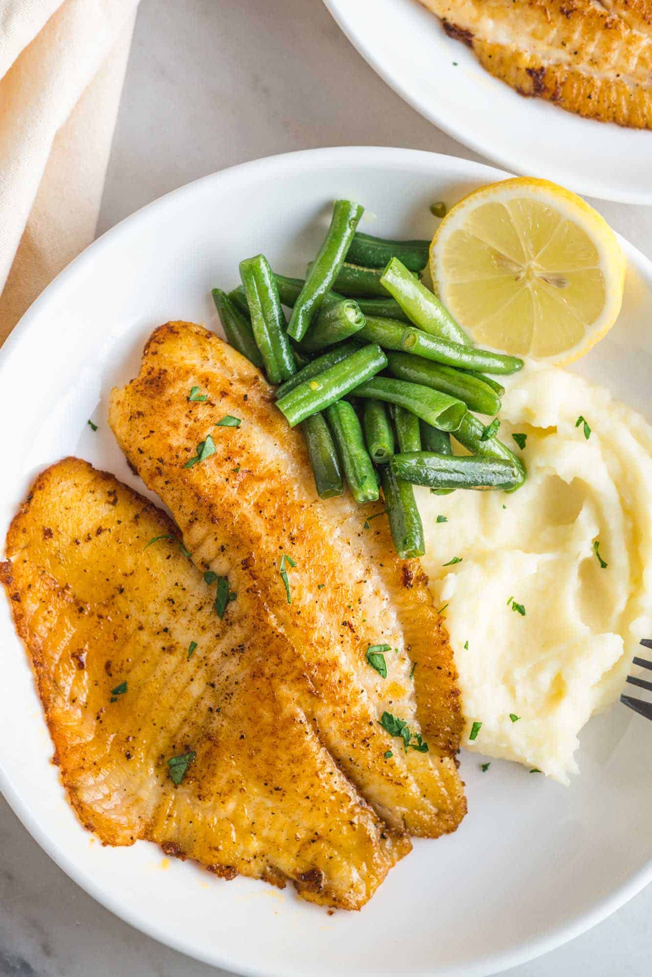 A white dinner plate filled with pan seared tilapia, green beans, mashed potatoes, and a half a lemon