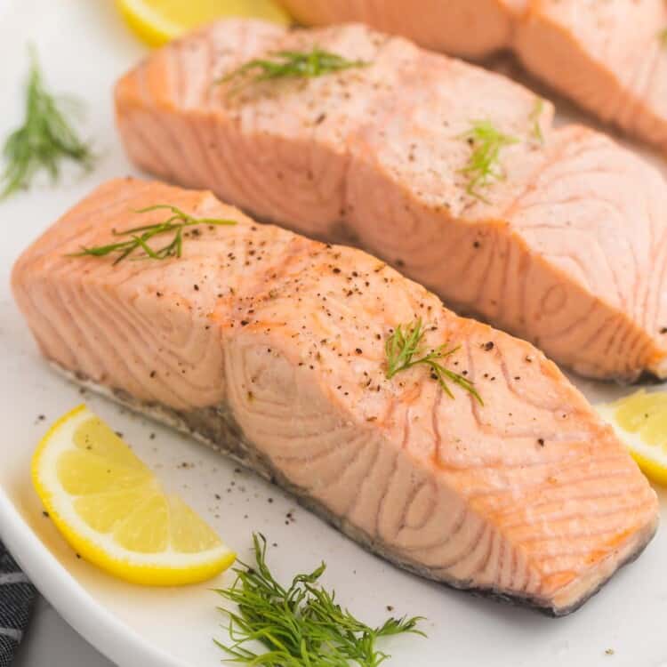 3 poached salmon fillets served on a white oval platter, with slices of lemon and fresh dill.