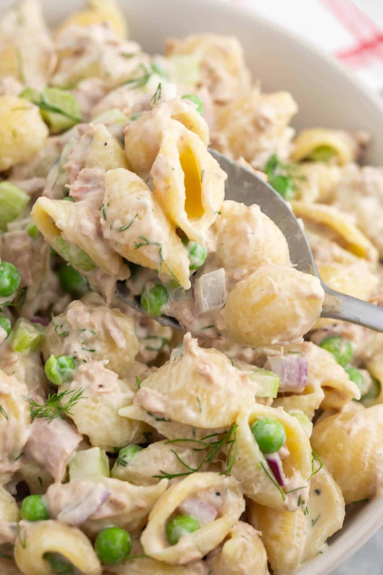 a closeup shot of a bowl of tuna macaroni salad made with shell pasta and peas being served with a metal spoon