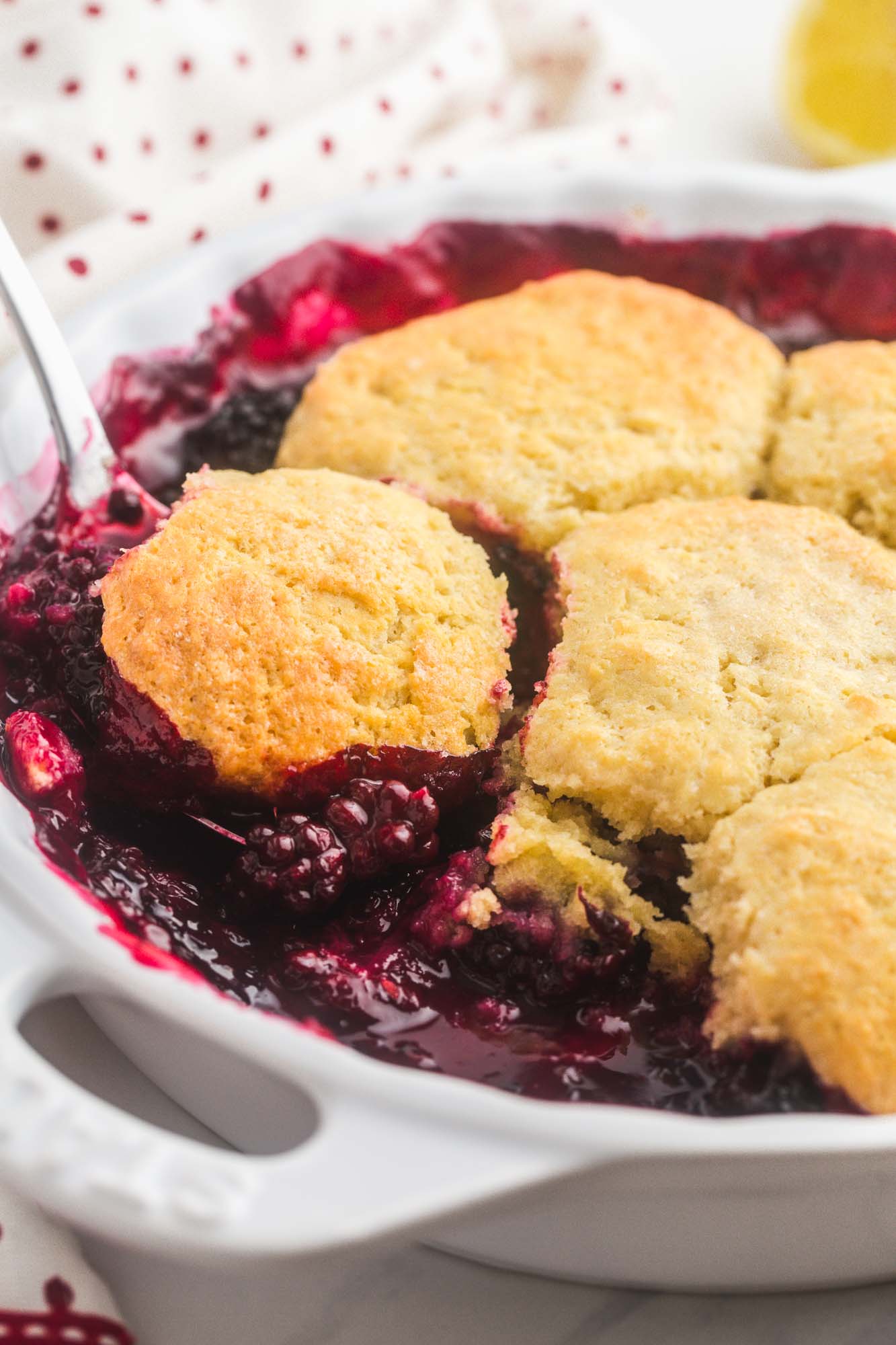 a round white baking dish of balckberry cobbler. A spoon is serving a scoop.