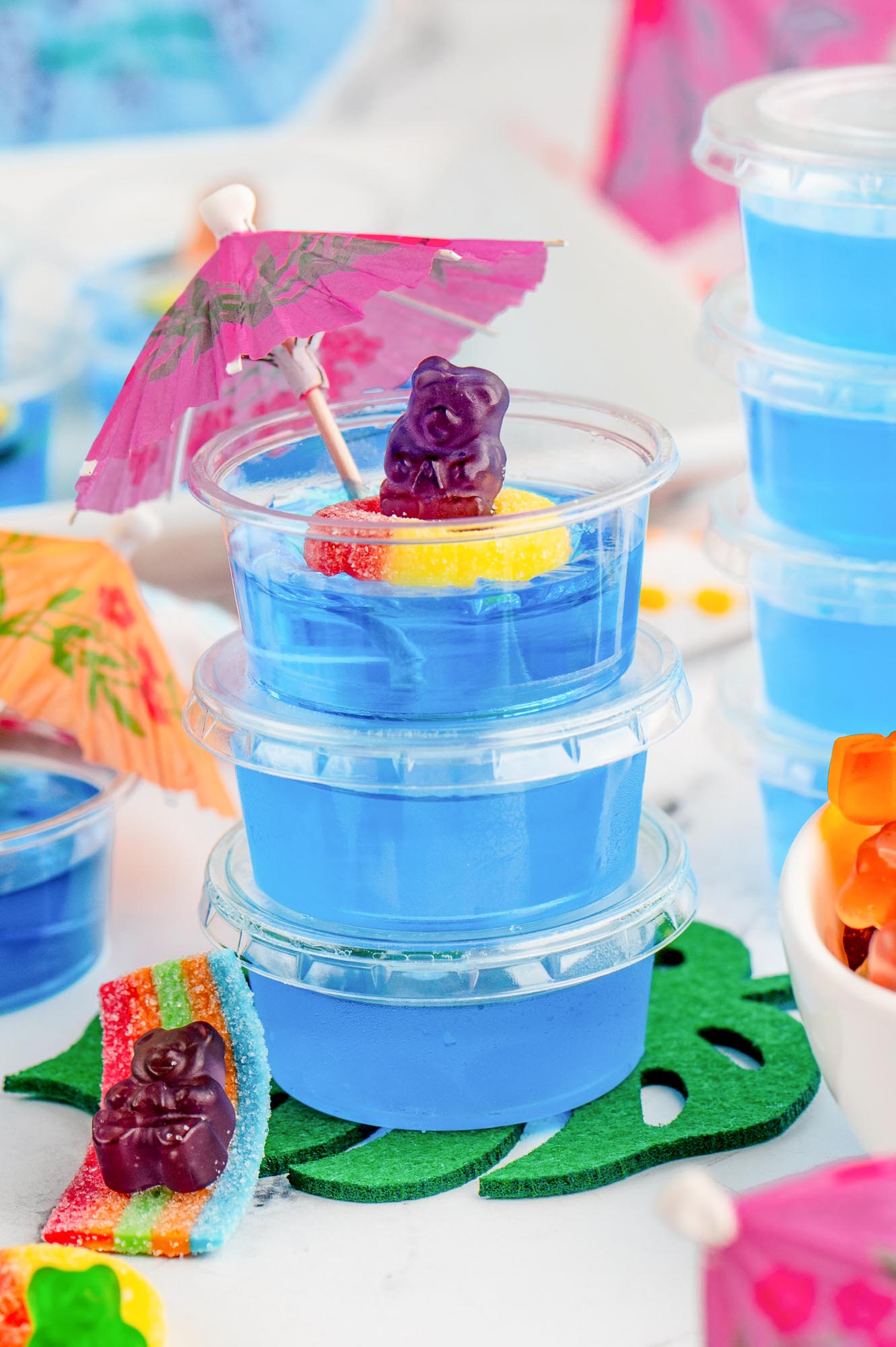Three blue pool party jello shots stacked, the top one has a gummy bear holding an umbrella on it.