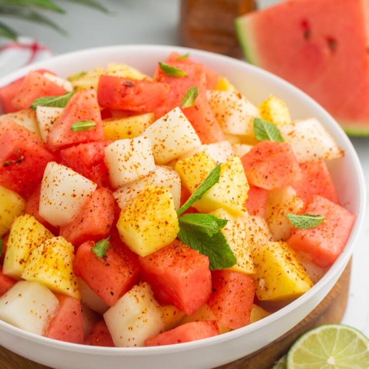 A bowl of Mexican fruit salad garnished with tajin, mint leaves, and lime.