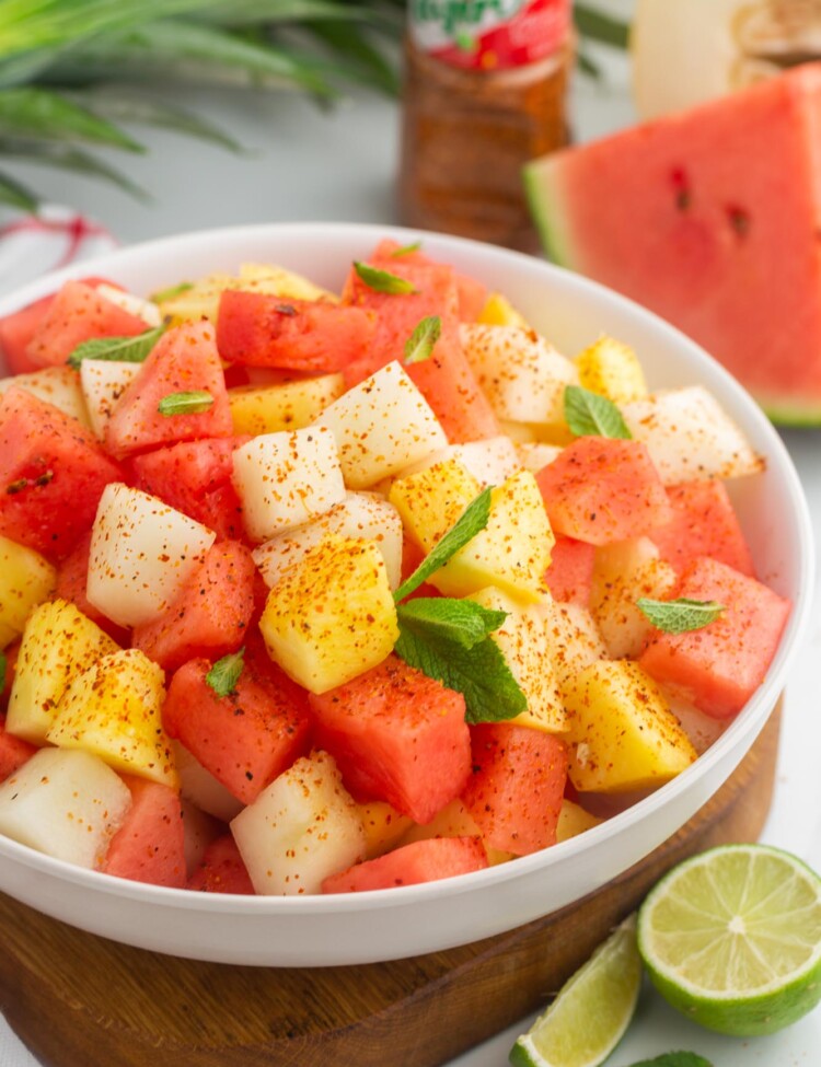 A bowl of Mexican fruit salad garnished with tajin, mint leaves, and lime.
