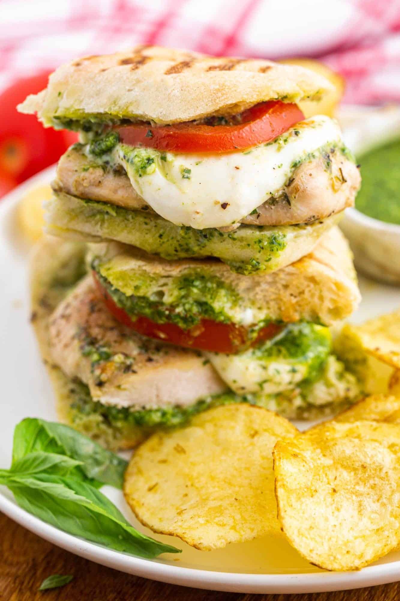 a plate with a chicken pesto sandwich, cut in half and stacked, next to some potato chips