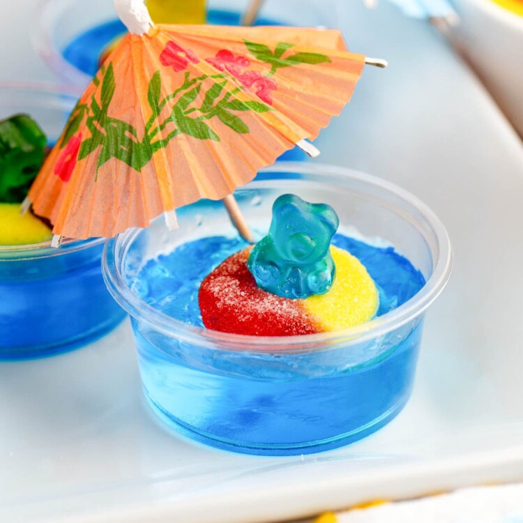 a blue jello shot with a blue gummy bear in a peach ring inner tube. A cocktail umbrella is stuck in the jello.