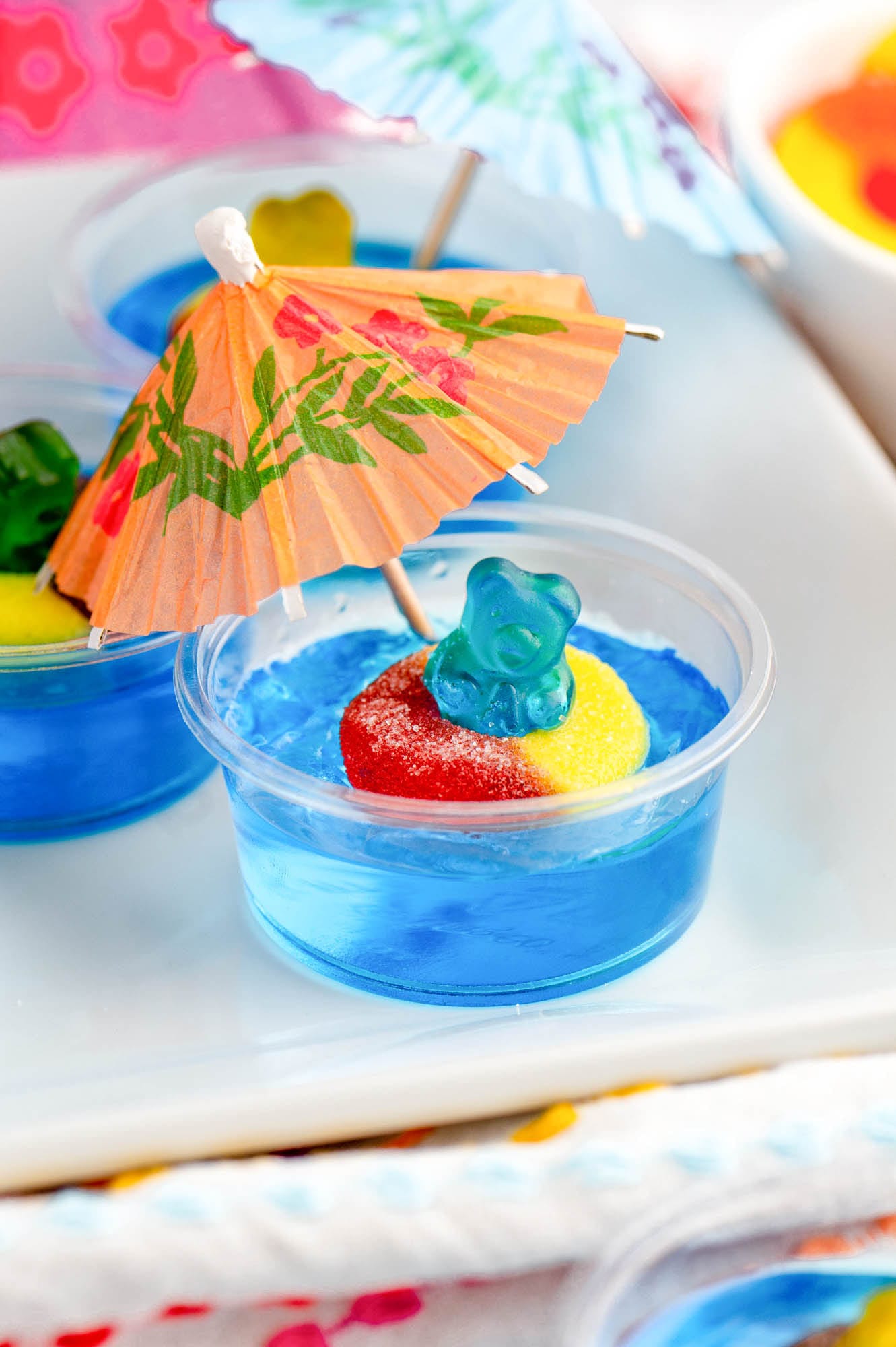 a blue jello shot with a blue gummy bear in a peach ring inner tube. A cocktail umbrella is stuck in the jello.