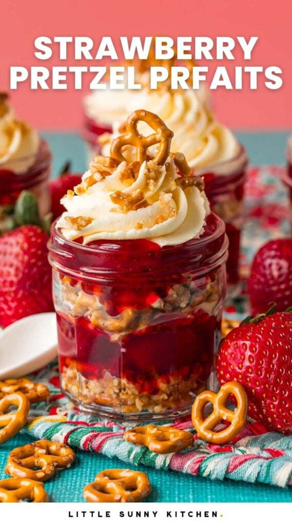 strawberry pretzel salad parfaits in small mason jars on a kitchen towel surrounded by fresh strawberries and mini pretzels