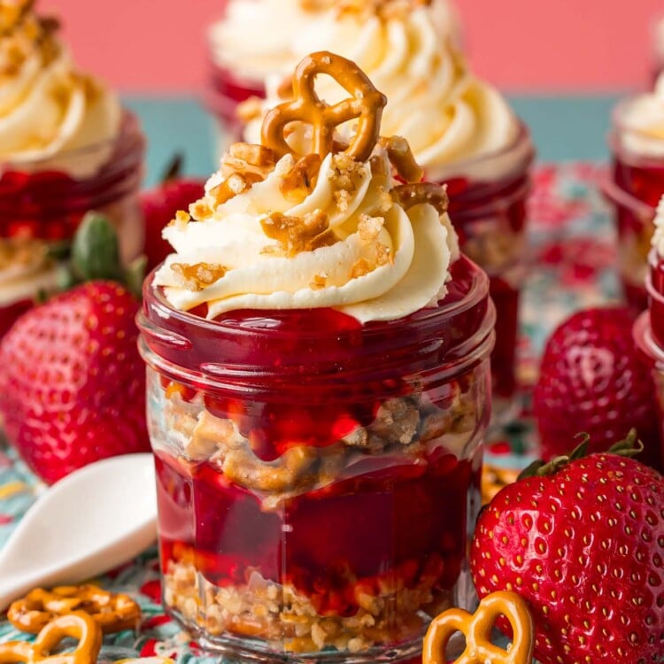 strawberry pretzel salad parfaits in small mason jars on a kitchen towel surrounded by fresh strawberries and mini pretzels