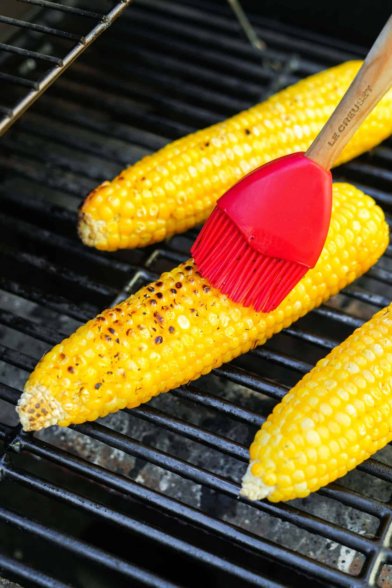 husked corn on an outdoor grill. A red silicone brush is adding butter.