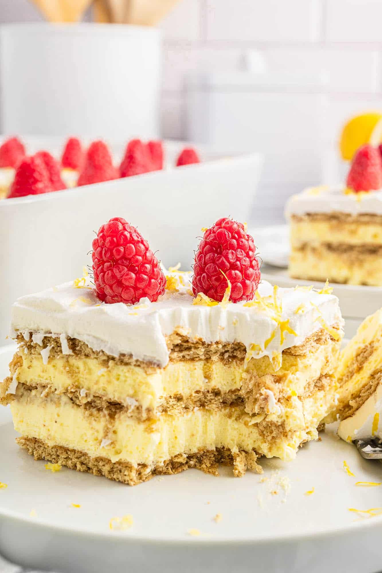 A square piece of graham cracker and Lemon pudding icebox cake with a bite taken off one corner, garnished with fresh raspberries and lemon zest.