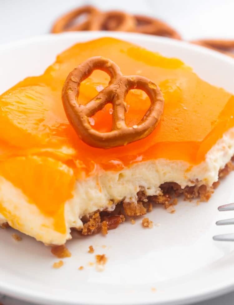 a square piece of orange jello pretzel salad on a plate, with bites taken. A pretzel is on top as garnish.